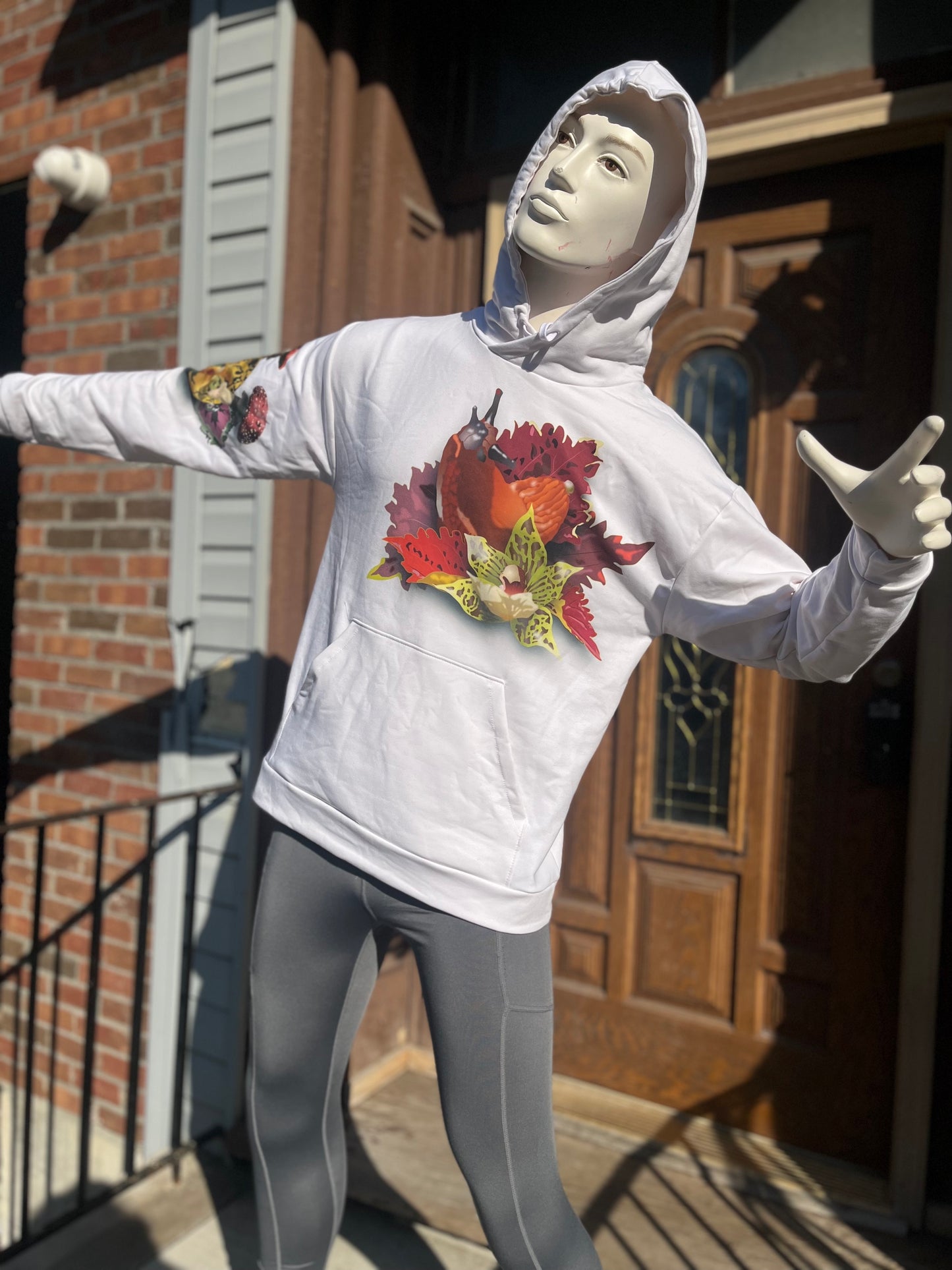 Mannequin wearing white hoodie showcasing an orange colored snail covered by a green orchid and red leaves.